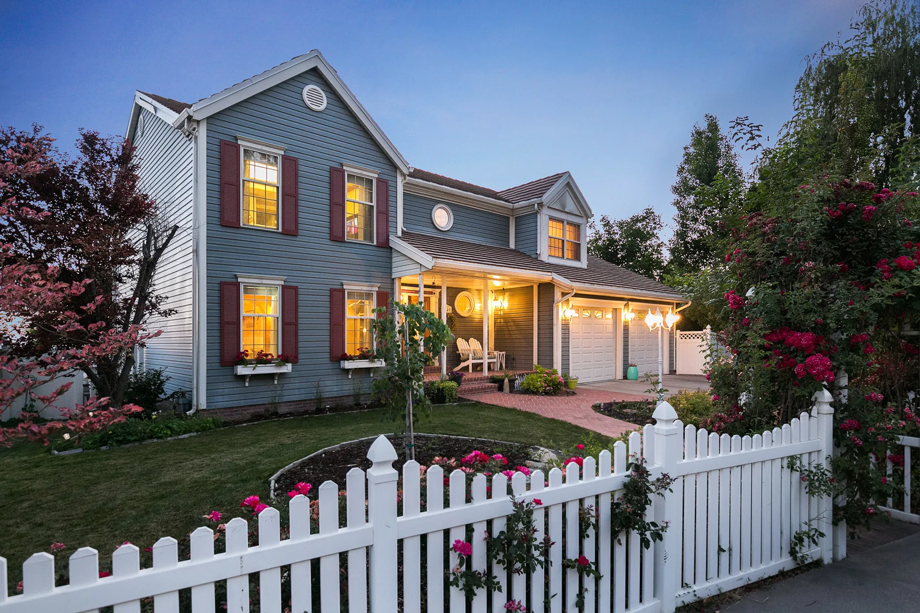 Blue two-story home illuminated at twilight behind a white picket fence
